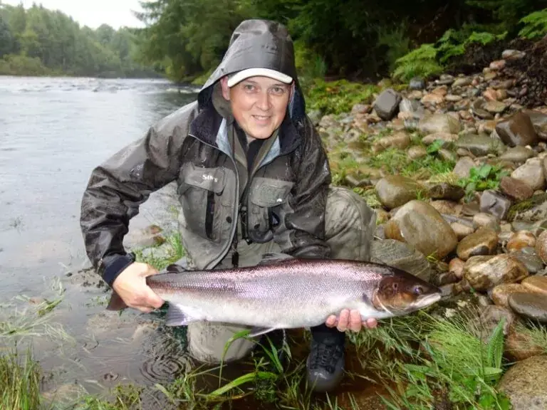 An angler in waterproofs kneels on a stony riverbank holding a large salmon in wet weather