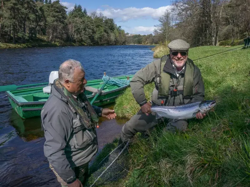 Two anglers beside a green boat, one kneeling on the bank holding a bright salmon