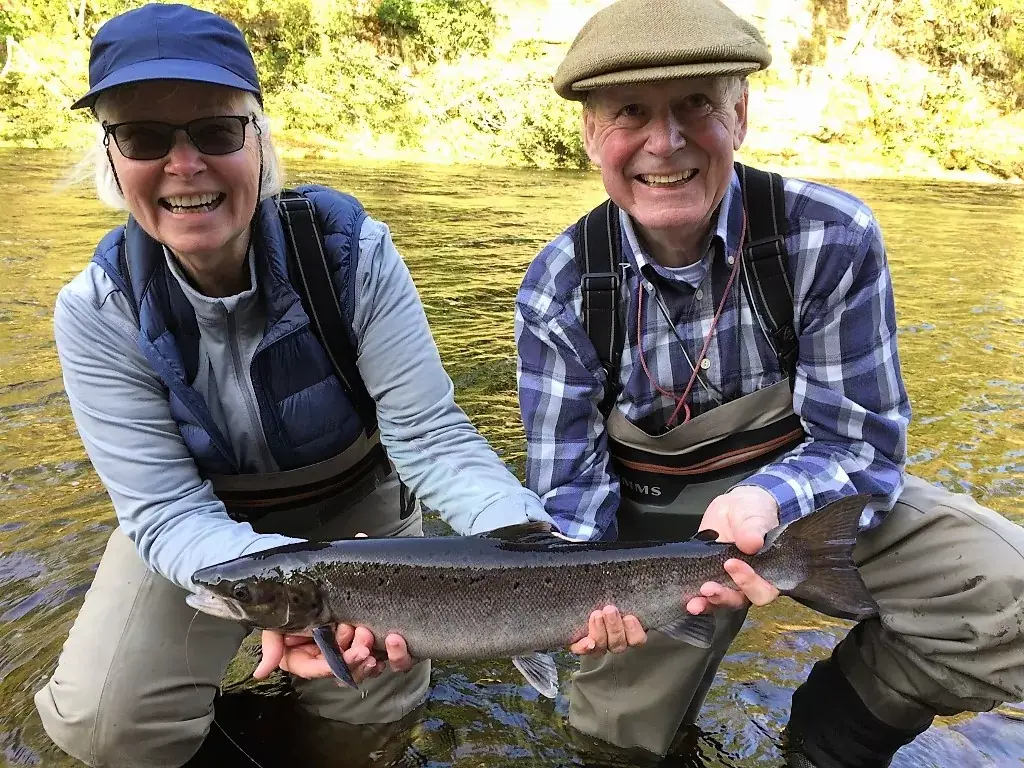 Two anglers in waders kneel in a river holding up a freshly caught salmon together