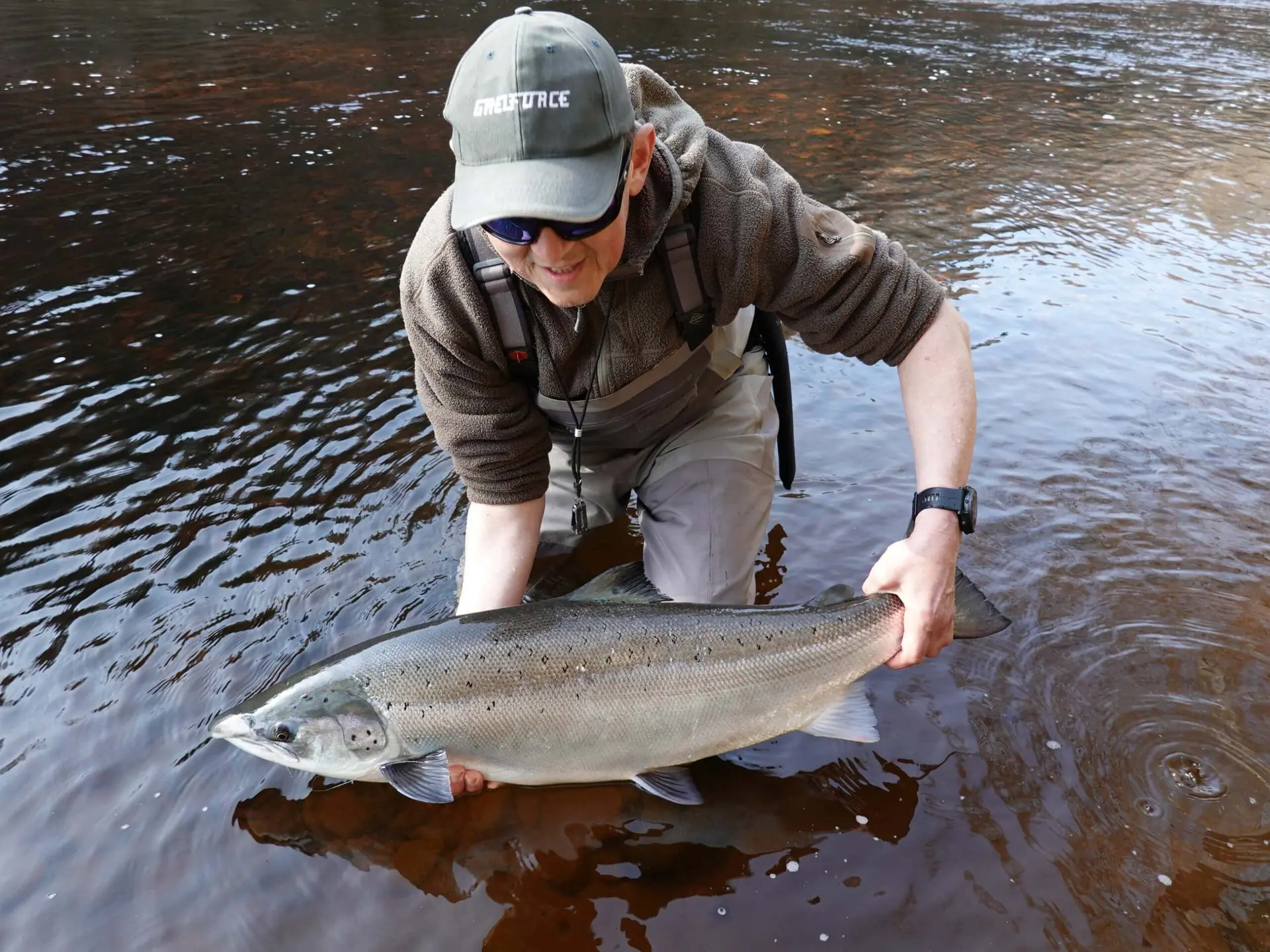 An angler kneeling in shallow river water holding a large salmon with both hands