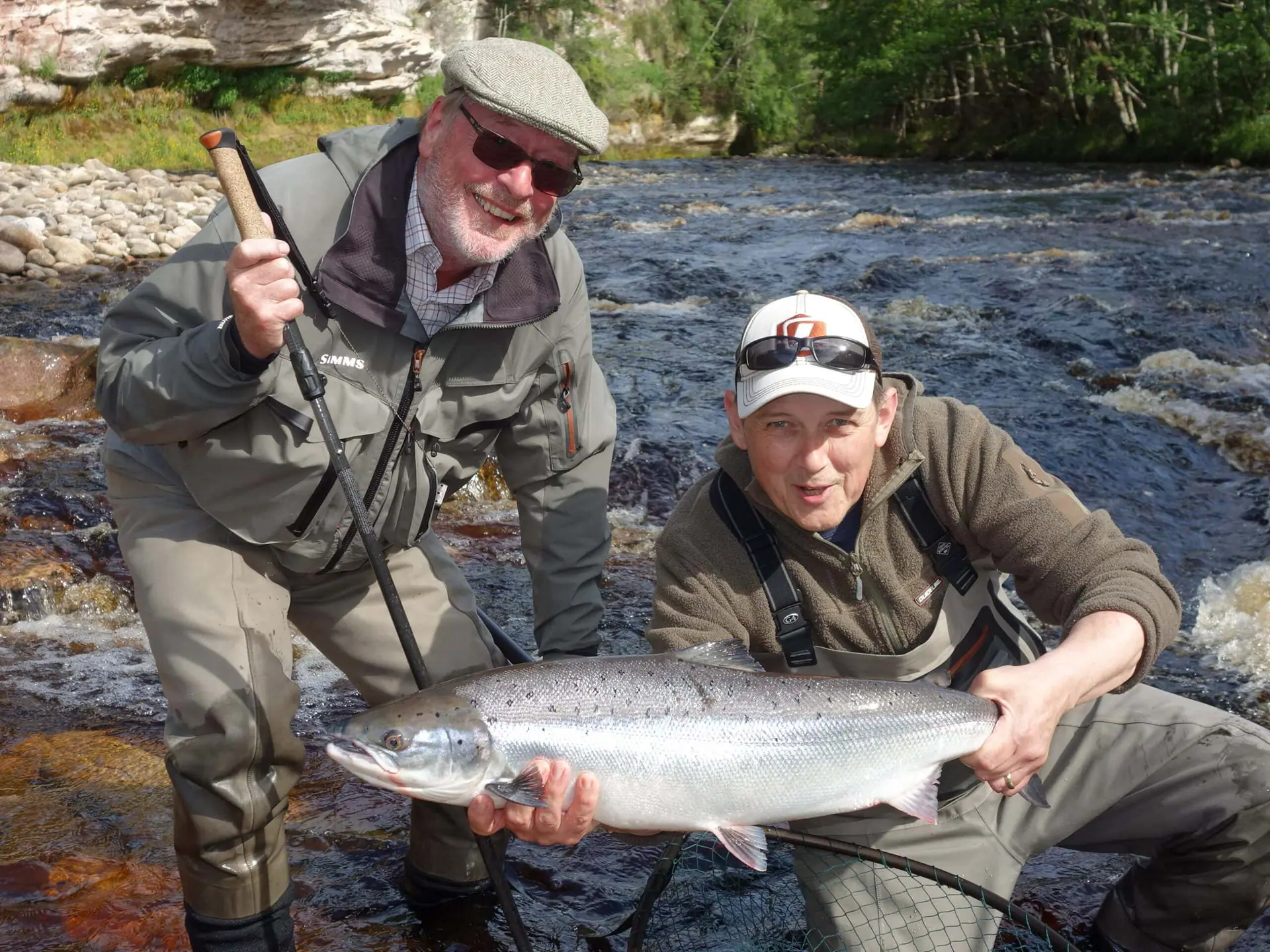 Two anglers kneeling in a rocky river holding a freshly caught salmon in sunshine