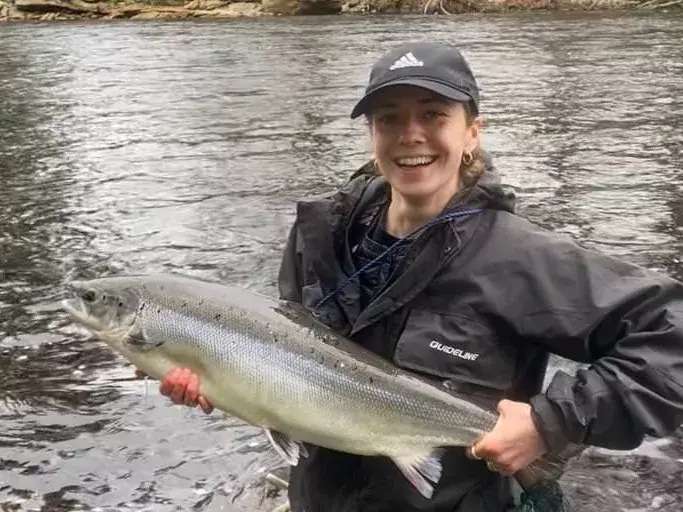 A smiling female angler in waders holds a large salmon beside the river