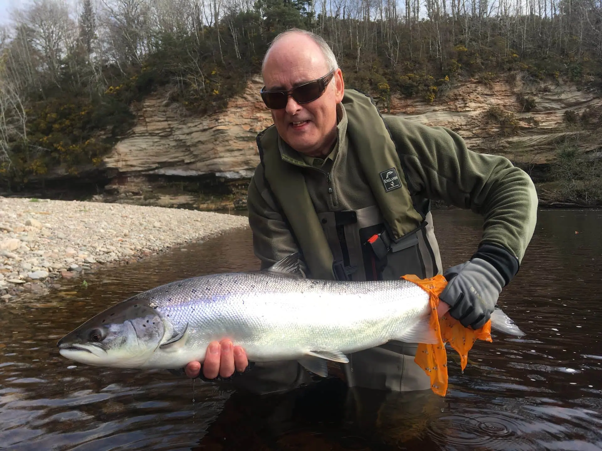 An angler wading the River Findhorn holds a bright salmon beside a steep wooded bank