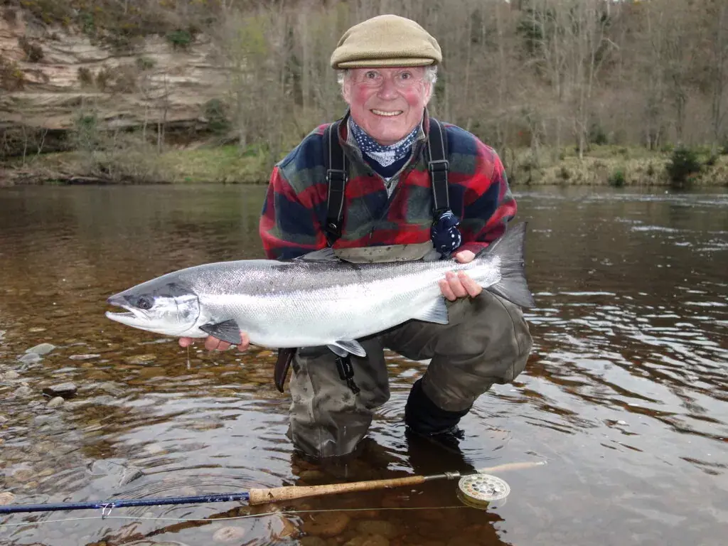 Ian Neale kneeling in the river holding a bright silver salmon, rod resting on the water