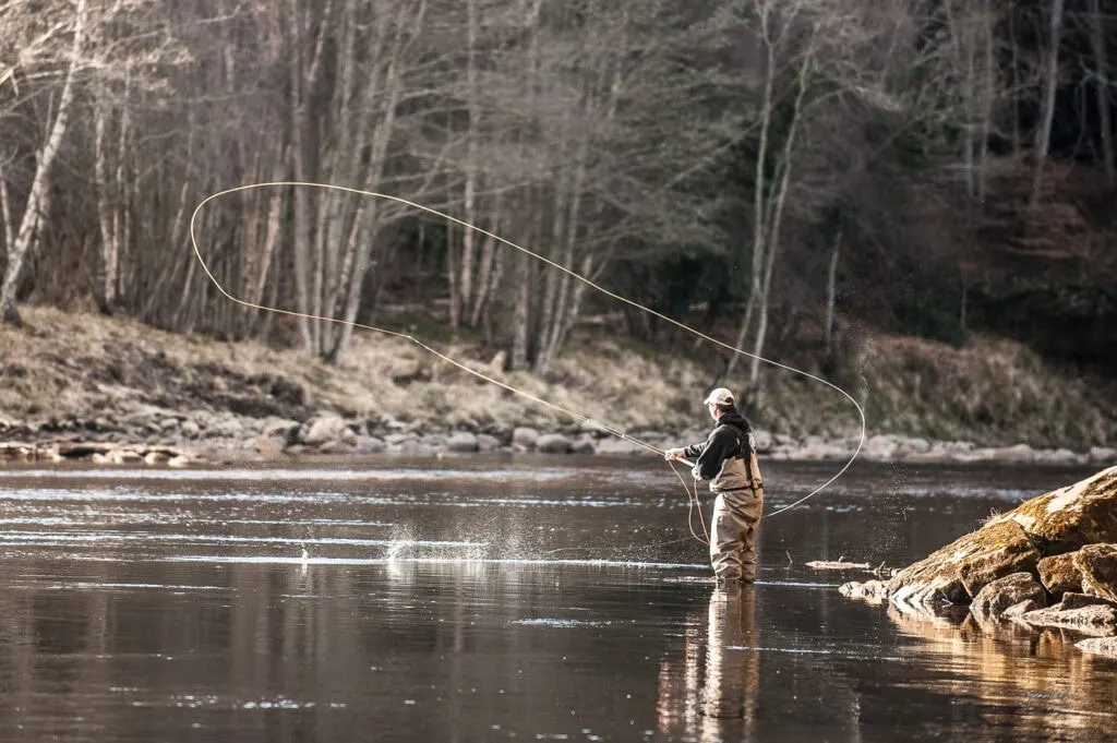 An angler Spey casting a wide loop of fly line on a river beside bare winter trees