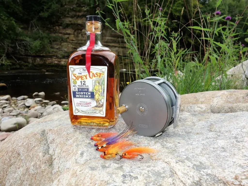 Whisky bottle, fly reel and orange salmon flies arranged on a riverside boulder