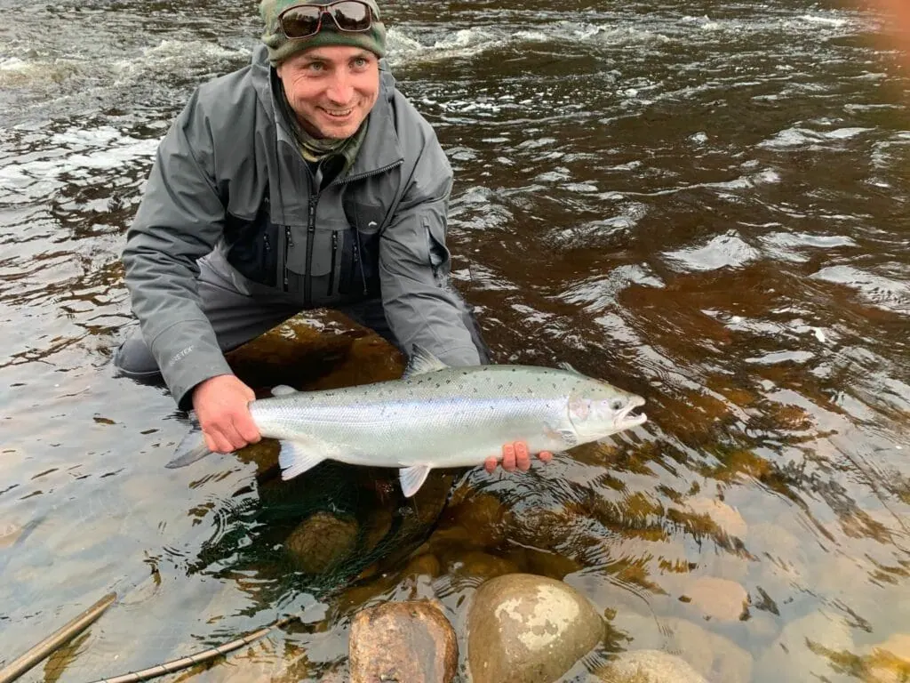 A guided client kneels in the river holding a bright silver salmon above rocky shallows