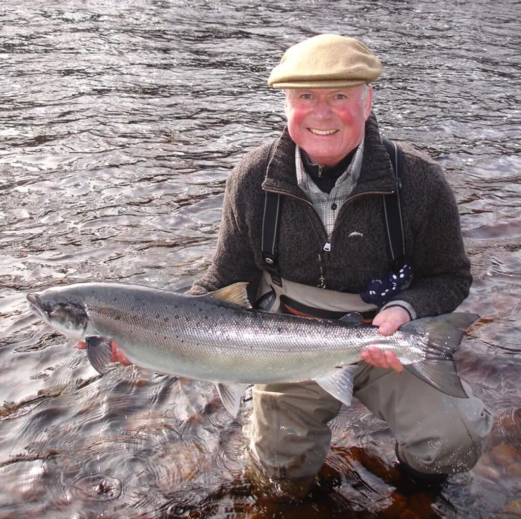 Ian Neale kneels in the river holding a bright salmon, smiling in soft daylight
