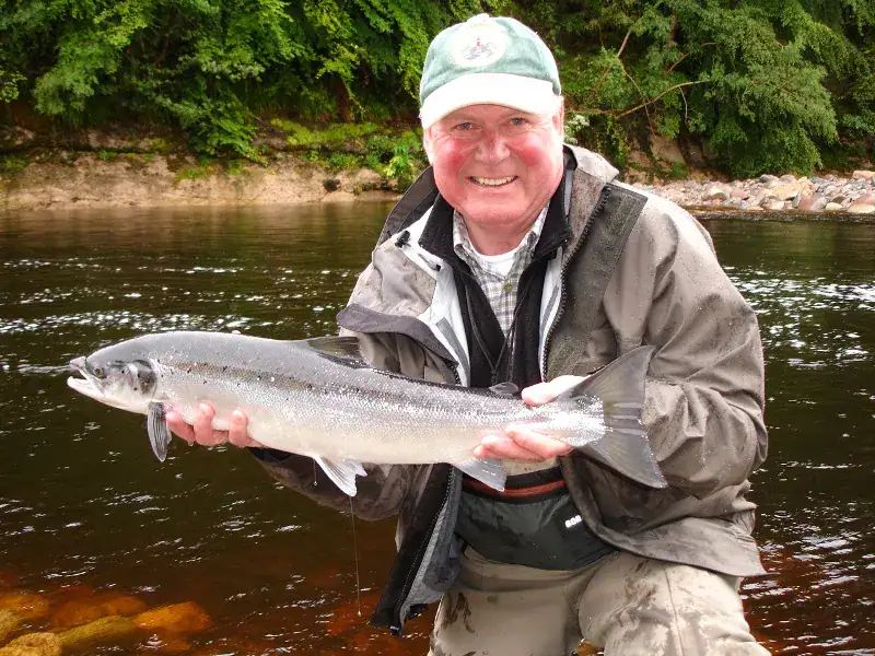 A smiling angler in waders holds a fresh salmon above a wooded river before release