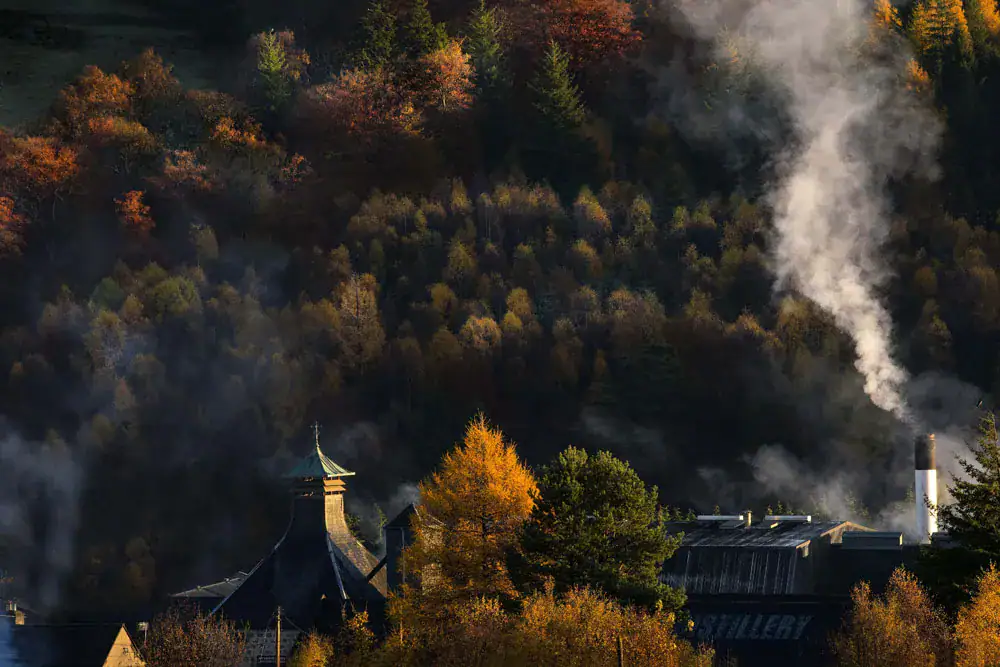 Balvenie Distillery pagoda roof and chimney smoke against an autumn wooded hillside in Speyside