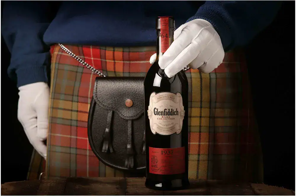 Gloved hand presenting a rare 1937 Glenfiddich whisky bottle against a tartan kilt and sporran